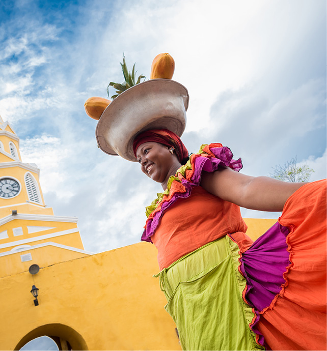 Local Palenquera woman wearing a colourful dress and a headpiece selling fruit next to a pretty yellow church